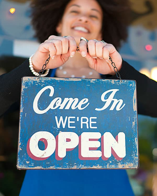 A woman smiles while holding a sign which says come in we’re open