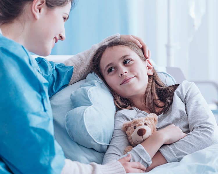 A young girl lays in bed with a teddy bear as her mother comforts her