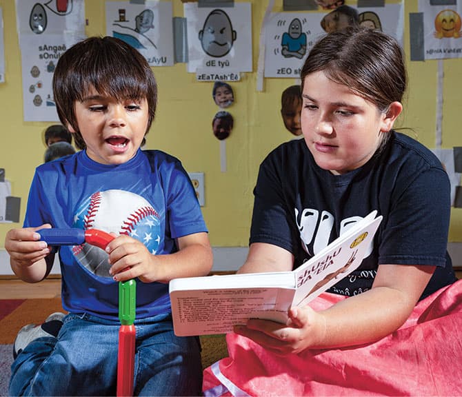 Enlargeable photo of an older kid teaching a younger one their Native language through reading