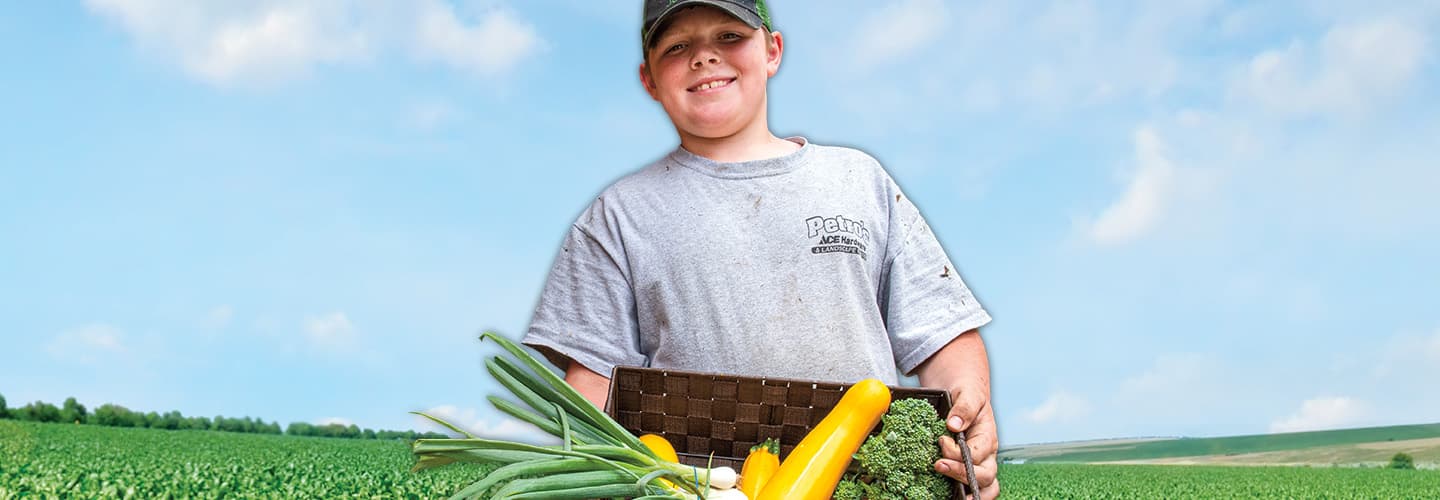 Kid holding a crate of fresh produce with a farm in the background