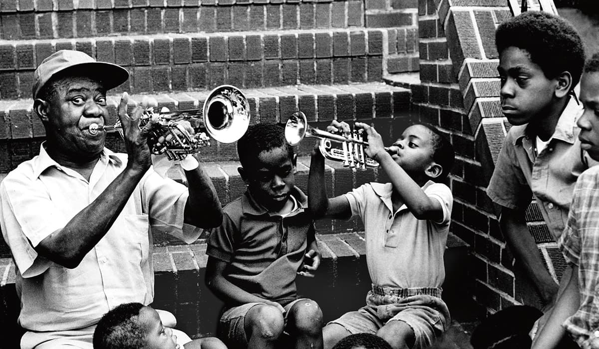 Black & white photo of Louis Armstrong playing trumpet with kids watching